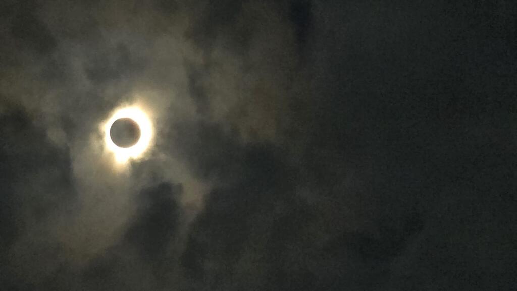A black disc with a glowing orange halo around it, shrouded by cloud in a dark sky.